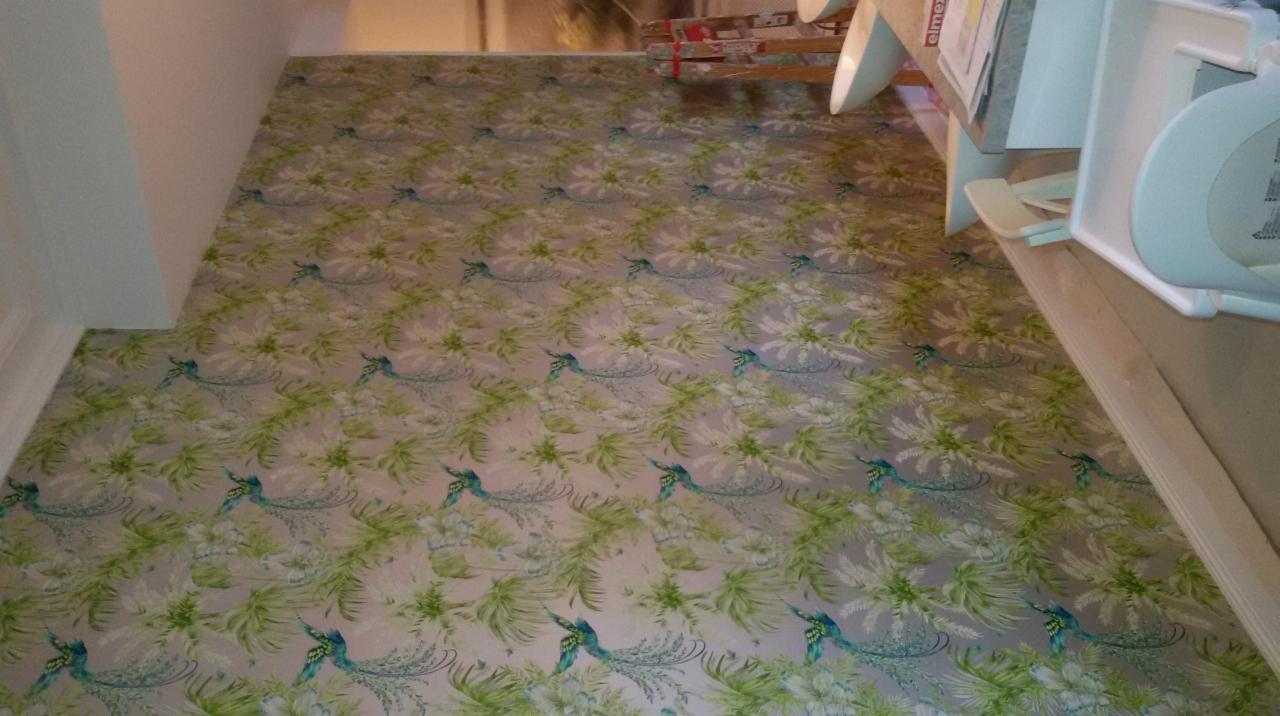 Podłoga wyłożona wzorzystą tapetą z motywem pawich piór w odcieniach zieleni i błękitu, widoczna krawędź ściany i fragment mebla.