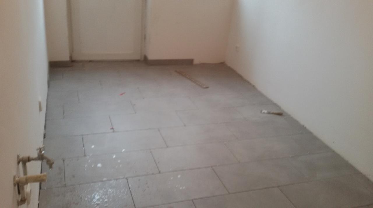 Ułożona szara terakota podłogowa w trakcie prac wykończeniowych, widoczne fragmenty ściany z instalacją hydrauliczną i listwa przypodłogowa.