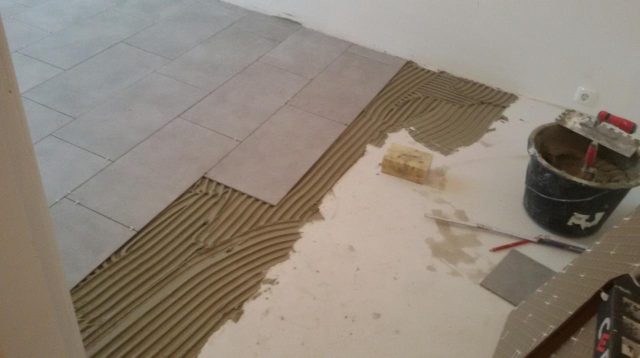 Układanie szarych płytek podłogowych na warstwie kleju, widoczny kubeł z zaprawą, poziomica i gąbka do czyszczenia.