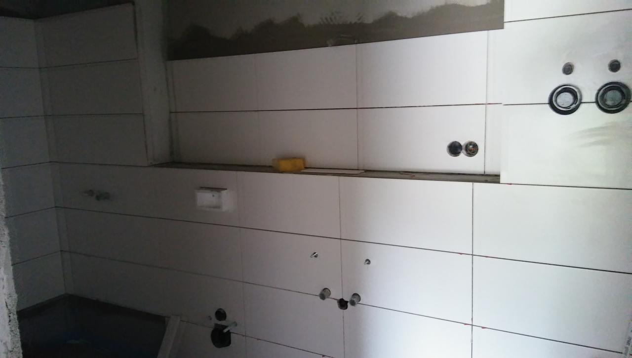 Białe płytki ścienne w trakcie montażu w łazience, widoczne otwory na instalacje i żółta gąbka na półce.