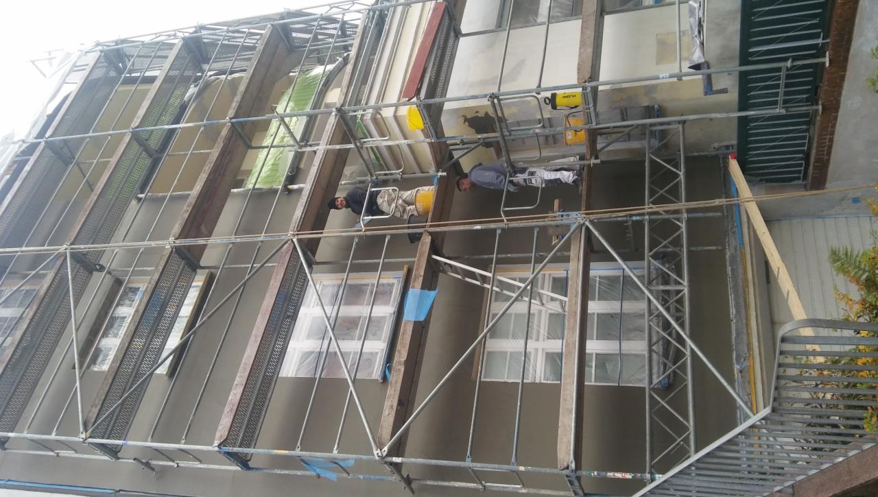 Dwóch pracowników na rusztowaniu podczas nakładania szarego tynku na elewację domu, okna zabezpieczone folią, widoczne narzędzia i żółte wiadra.