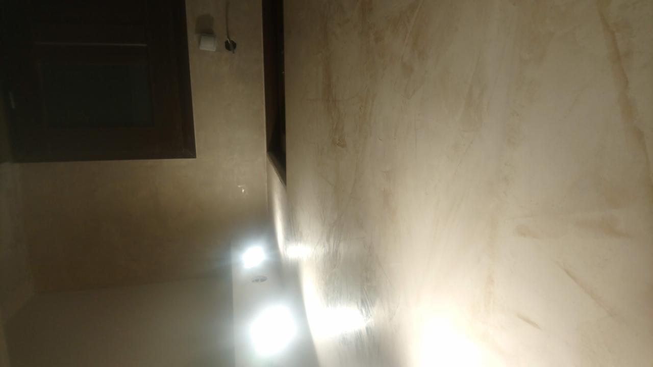 Ściana wewnętrzna z efektem dekoracyjnym w odcieniach beżu i brązu, widoczne ciemne obramowanie okna i punktowe oświetlenie sufitowe.