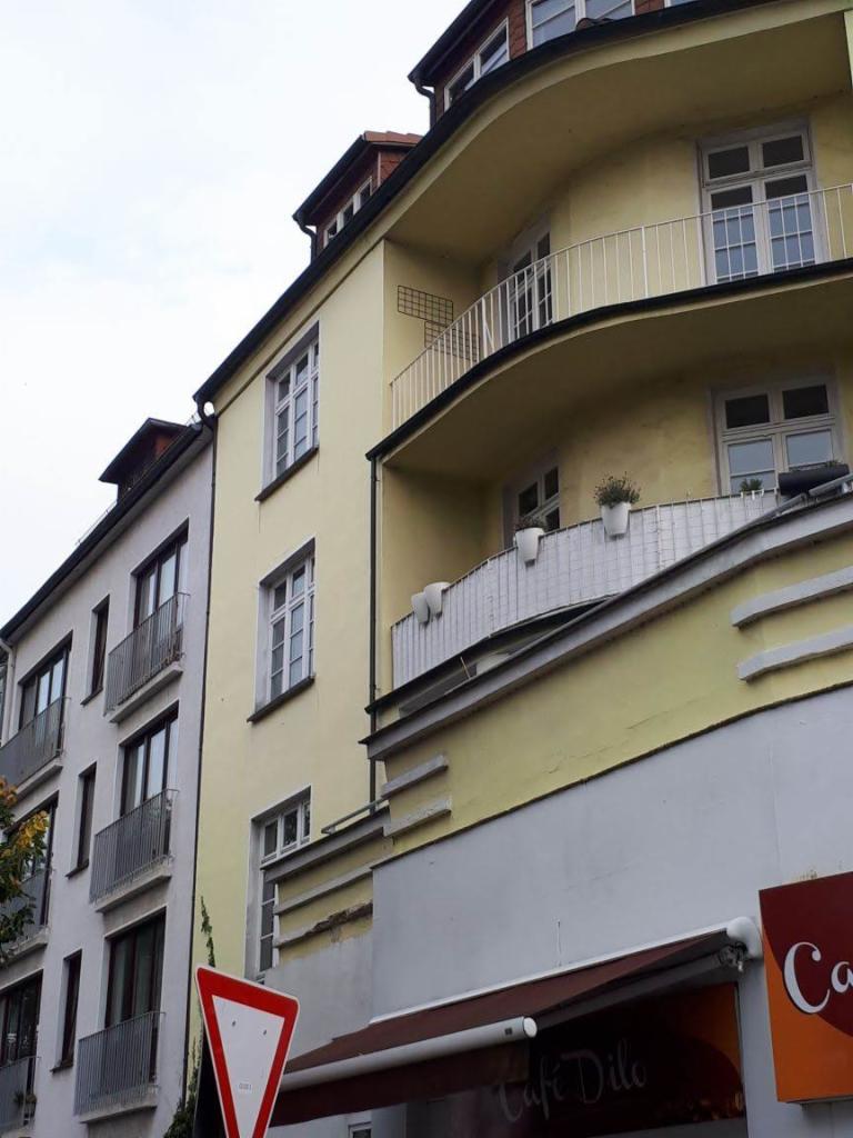 Żółta kamienica z zaokrąglonymi balkonami, widok z dołu, z widocznym fragmentem znaku drogowego i markizy kawiarni w dolnej części kadru.