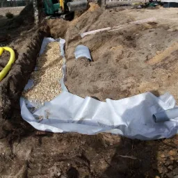 Wykop w ziemi wyłożony geowłókniną, częściowo wypełniony drobnym żwirem, z widocznymi rurami drenażowymi i fragmentem żółtego węża; w tle mała koparka.