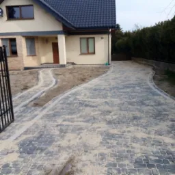 Ułożona, szara kostka brukowa przed domem z jasną elewacją i ciemnym dachem, z widocznymi liniami oddzielającymi ścieżki.