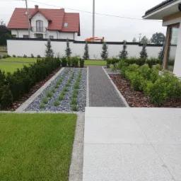 Nowoczesny ogród z równo przystrzyżonym trawnikiem, ścieżką z szarej kostki, rabatami z ozdobnymi kamieniami i roślinami, oraz białym murem w tle.