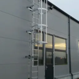 Zewnętrzna metalowa drabina z zabezpieczeniem przymocowana do ściany budynku z płyt warstwowych, prowadząca do dachu.