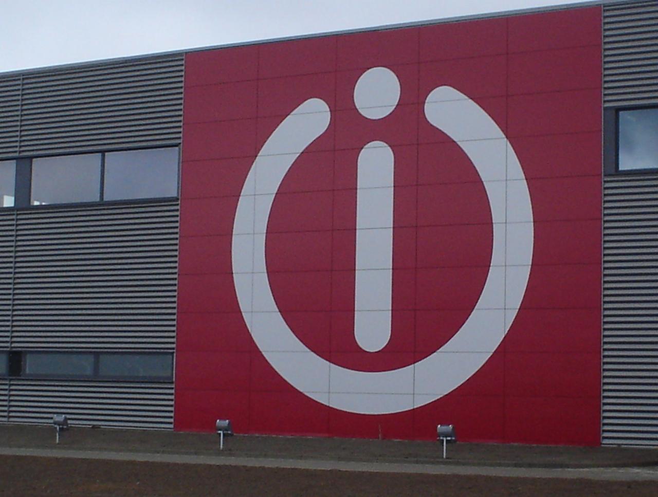Duże logo firmy odzieżowej na fasadzie budynku z szarej blachy falistej i czerwonych paneli. Logo w kolorze białym na czerwonym tle, przedstawia stylizowaną literę 'i' otoczoną okręgiem z przerwą u...