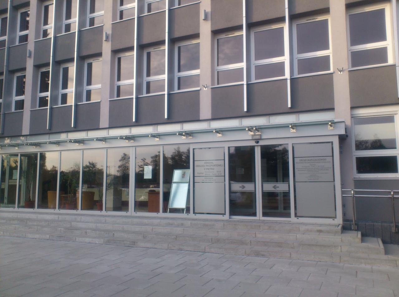 Elewacja budynku z szarymi ścianami, rzędem okien i szklanym wejściem z informacjami na szybach