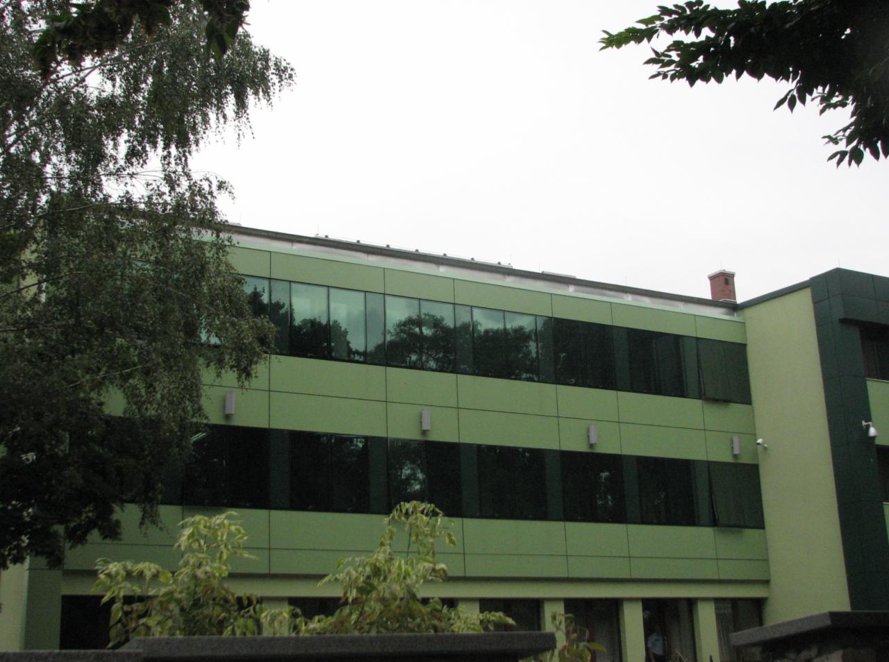 Dwukondygnacyjny budynek biurowy w odcieniach zieleni z dużymi oknami, częściowo zasłonięty drzewami i krzewami.