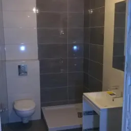 Wykończona łazienka z szarymi i białymi płytkami, kabiną prysznicową bez brodzika, wiszącą toaletą i umywalką z szafką, widok z perspektywy wejścia.