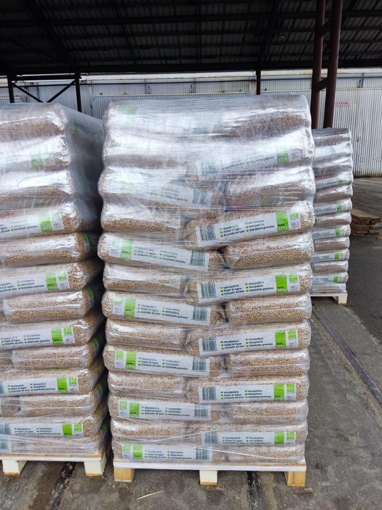 Ułożone palety z workami pelletu drzewnego, owinięte folią, z widocznymi etykietami informacyjnymi. Magazyn zadaszony w tle.