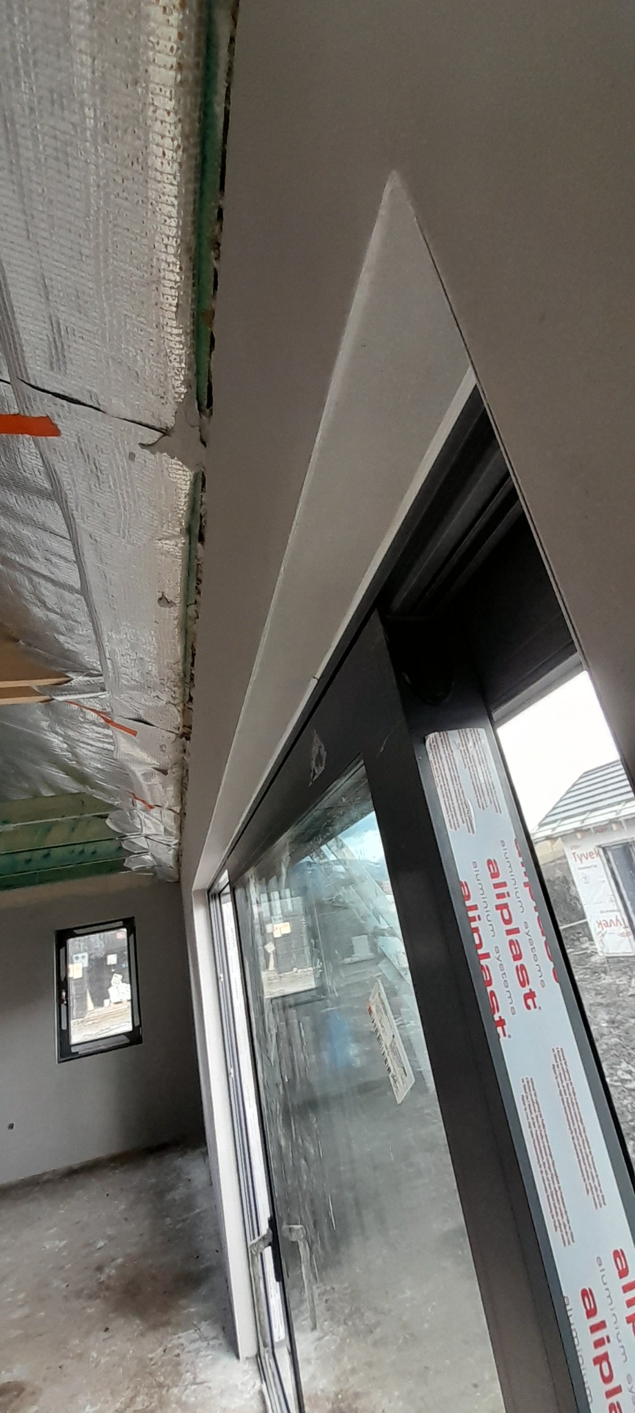 Wnętrze w trakcie prac wykończeniowych: widok na okno z naklejką producenta, sufit z izolacją, ściany po nałożeniu gładzi, małe okno w oddali.