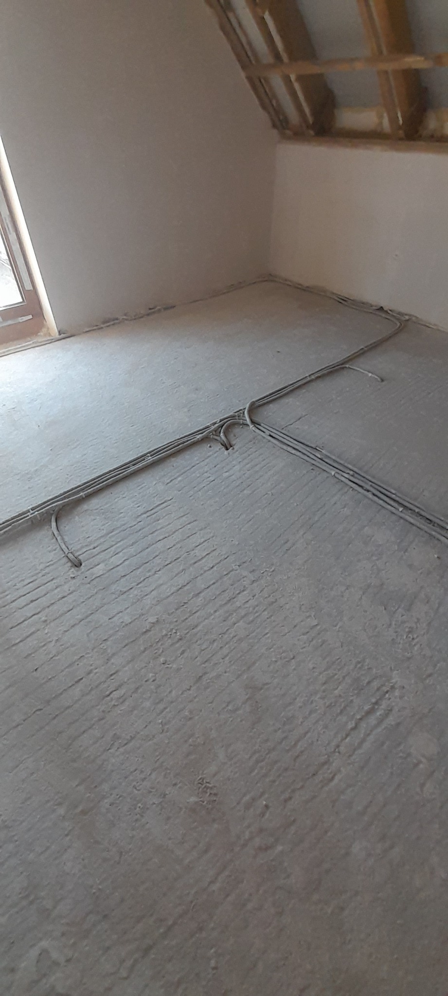 Surowe wnętrze poddasza z widoczną instalacją elektryczną na posadzce przed wylewką. Ściany w trakcie wykańczania, widoczna konstrukcja drewniana dachu.