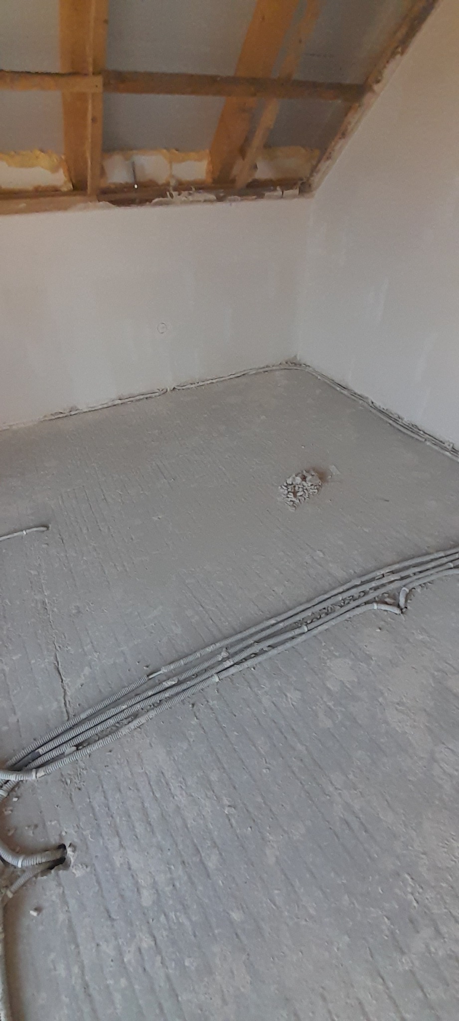 Surowe wnętrze w trakcie prac wykończeniowych: ściany pokryte jasnym tynkiem, widoczne przewody elektryczne w peszlach na podłodze z wylewką, drewniany szkielet stropu z izolacją z pianki.