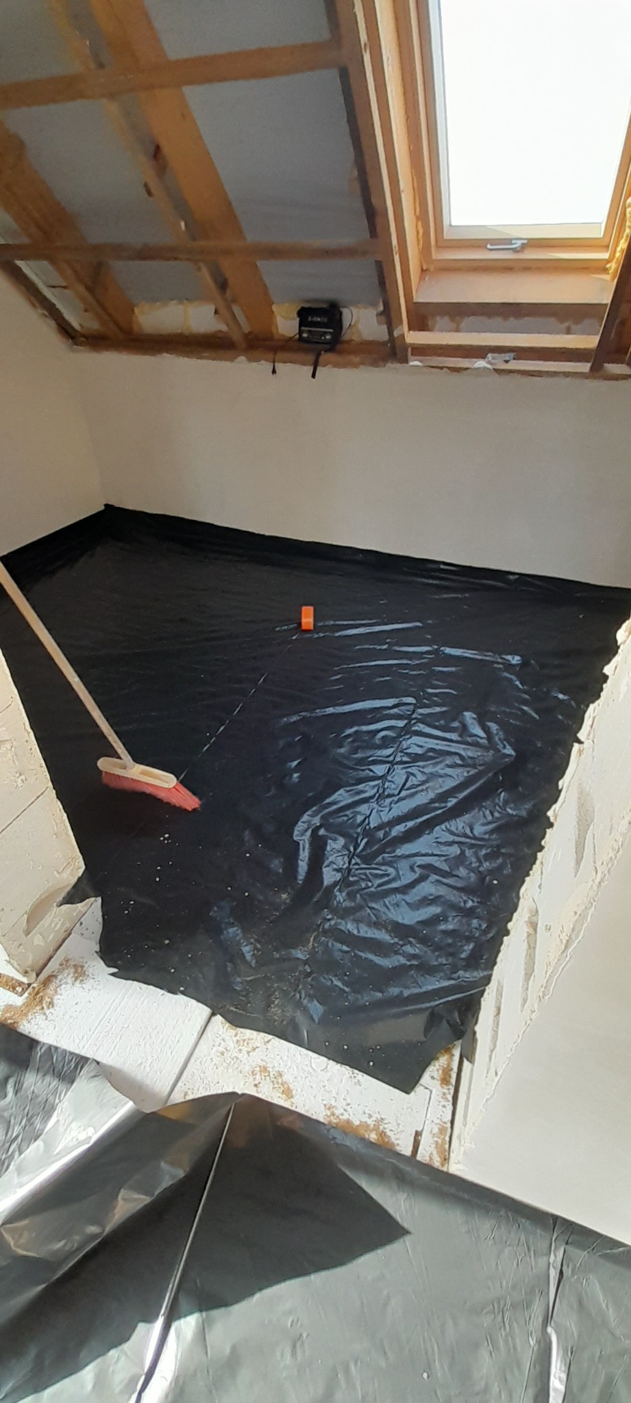 Przygotowanie pomieszczenia do prac wykończeniowych: podłoga zabezpieczona czarną folią, widoczny fragment sufitu z oknem dachowym i elementami konstrukcyjnymi, ściany wstępnie wykończone, leżąca...