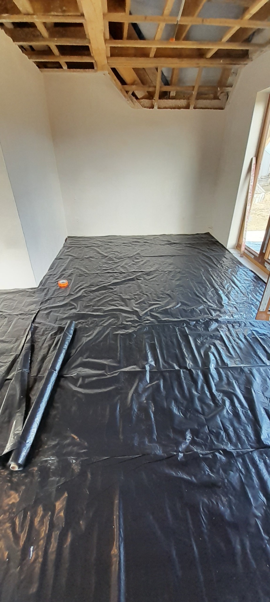 Przygotowanie do prac wykończeniowych w pomieszczeniu: podłoga zabezpieczona czarną folią, widoczny fragment drewnianej konstrukcji sufitu, ściany przygotowane do gładzi.