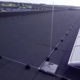 System ochrony odgromowej z przewodami i masztami na płaskim, bitumicznym dachu budynku przemysłowego z widocznym świetlikiem dachowym.