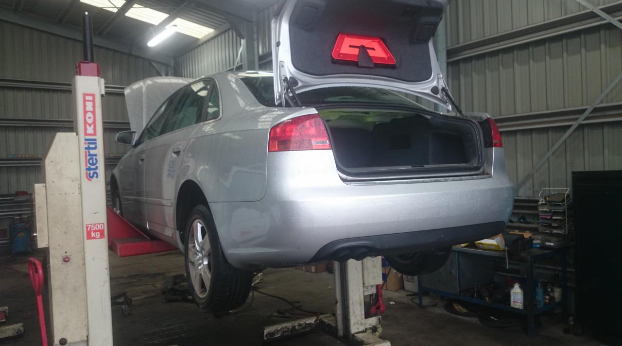 Srebrny samochód na podnośniku w warsztacie, otwarty bagażnik z trójkątem ostrzegawczym, podniesiona maska, widoczny podnośnik Stertil Koni.