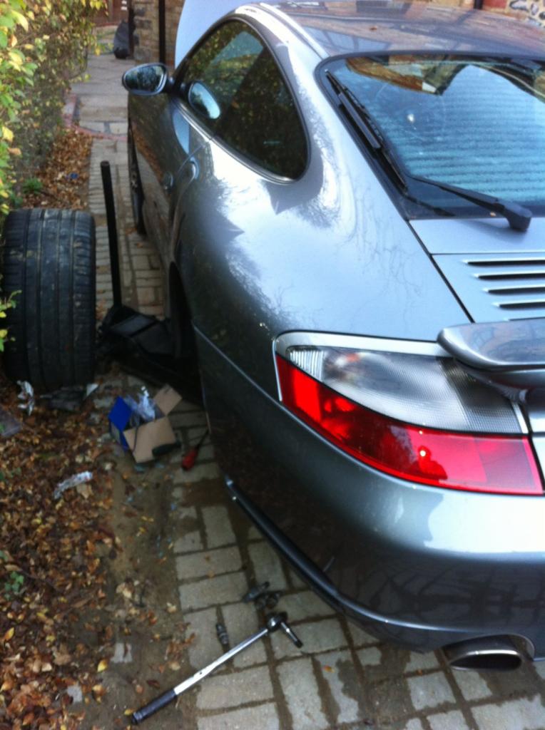 Szary samochód sportowy z otwartą maską, podnośnikiem i kluczem leżącym obok, prawdopodobnie podczas wymiany opony na podjeździe z kostki brukowej.