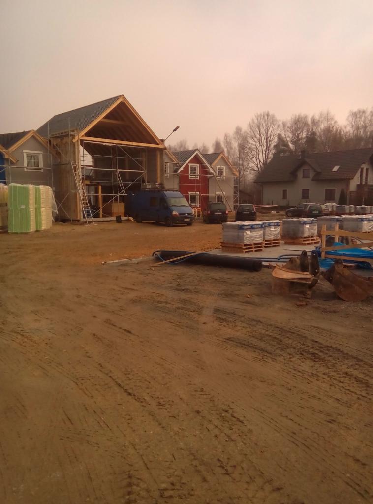 Budowa domów jednorodzinnych w trakcie realizacji, widoczne rusztowania, materiały budowlane, samochód dostawczy i wykopane fundamenty.