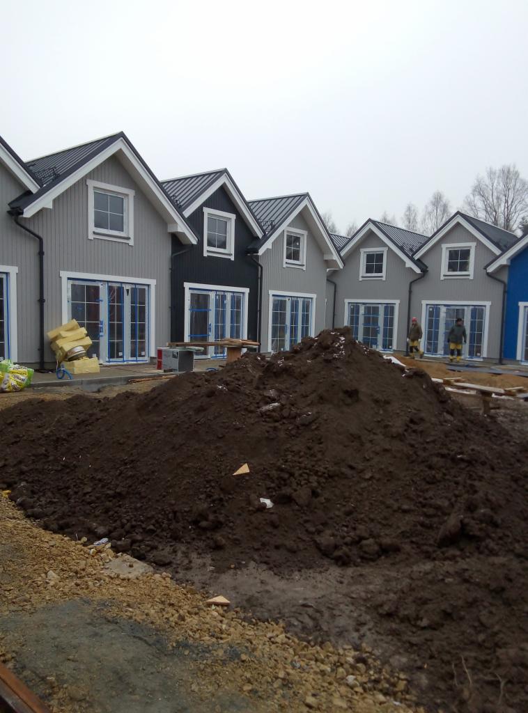 Rząd szarych domów jednorodzinnych z ciemnymi dachami w trakcie budowy, widoczny stos ziemi na pierwszym planie i robotnicy w oddali.