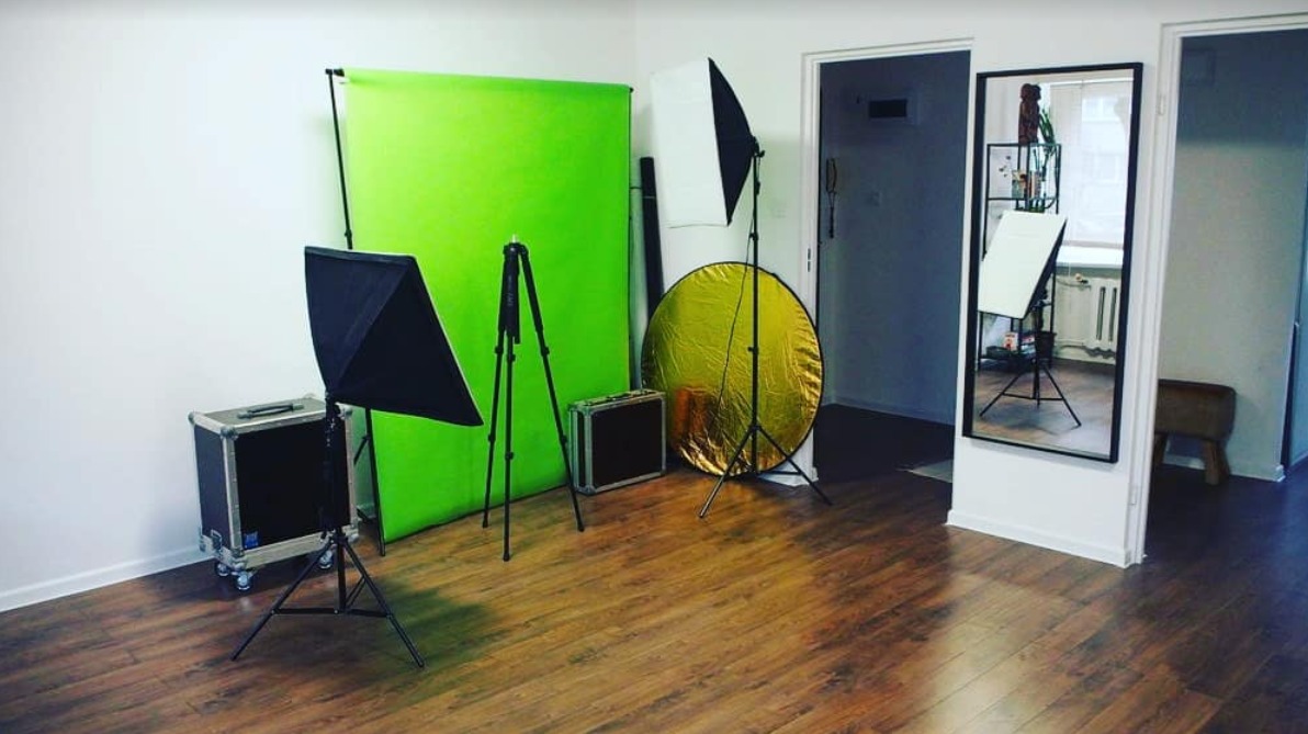 Studio fotograficzne z zielonym tłem, lampami studyjnymi, softboxem i złotą blendą, widoczne drewniane panele podłogowe i lustro z odbiciem sprzętu.