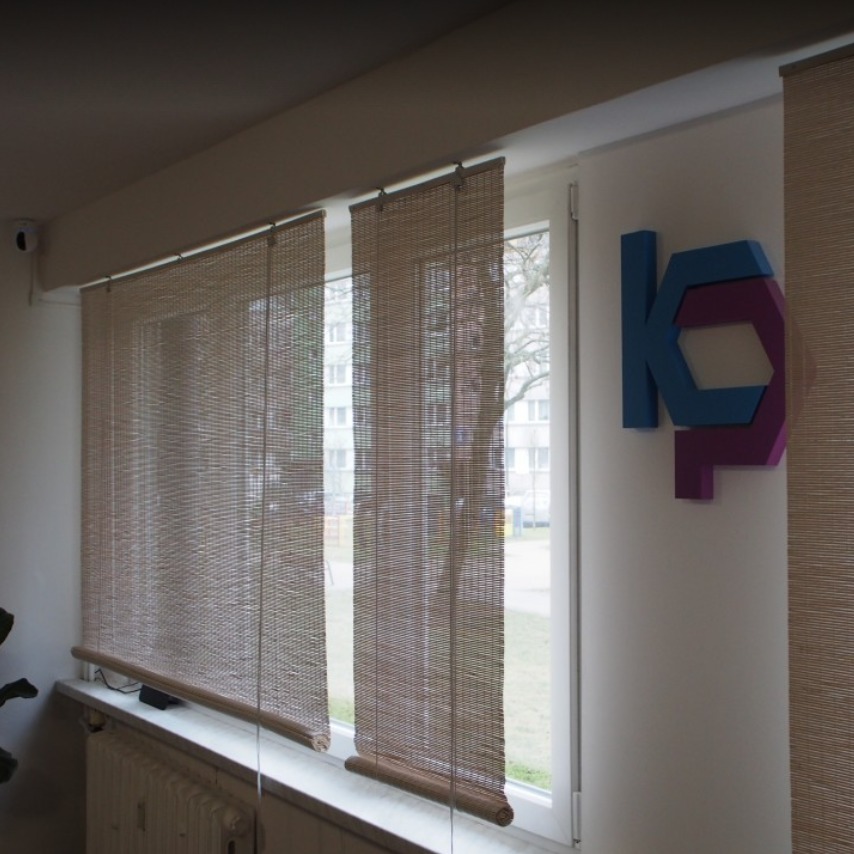 Wnętrze biura z bambusowymi roletami, logo firmy w odcieniach niebieskiego i fioletu na ścianie, widok z okna na budynek mieszkalny.