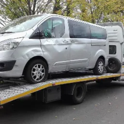 Uszkodzony srebrny Ford Transit na platformie lawety na tle jesiennego krajobrazu. Widoczne wgniecenia karoserii po stronie kierowcy i pęknięta opona.