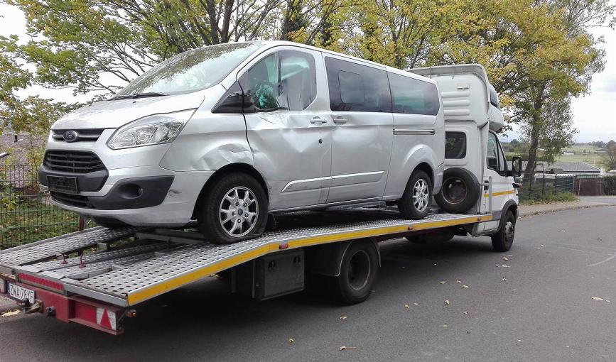 Uszkodzony srebrny Ford Transit na platformie lawety na tle jesiennego krajobrazu. Widoczne wgniecenia karoserii po stronie kierowcy i pęknięta opona.