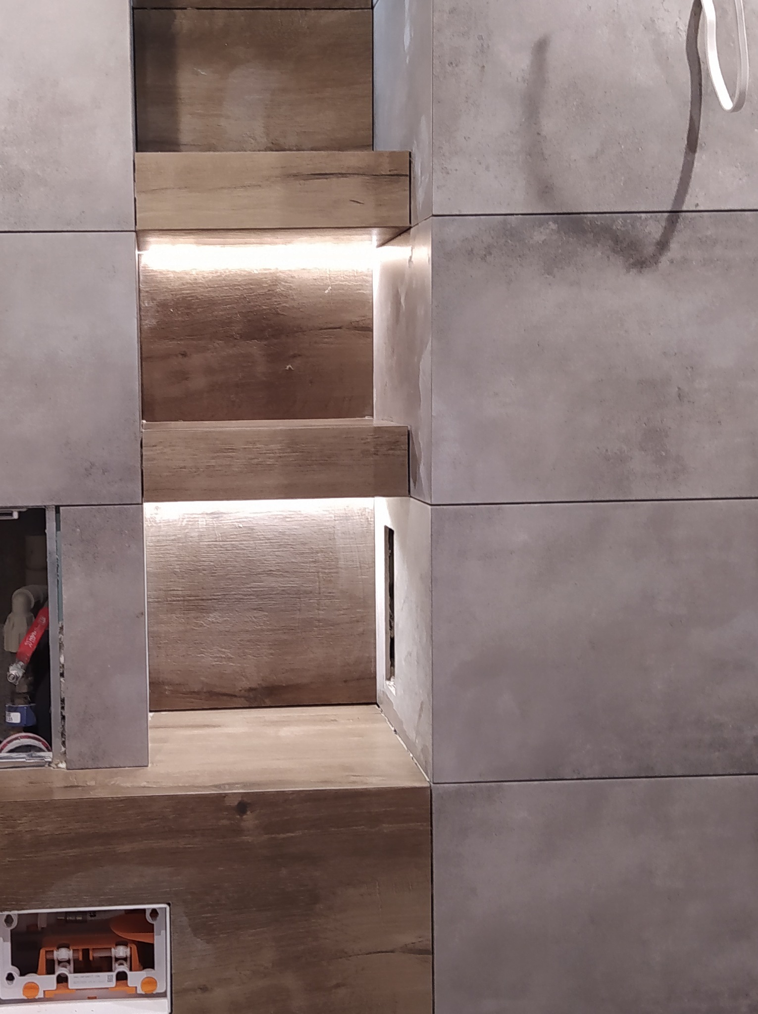 Narożna półka z drewnianymi wstawkami i podświetleniem LED, wkomponowana w szare płytki ścienne, widoczne elementy instalacji wodnej i elektrycznej w ścianie.