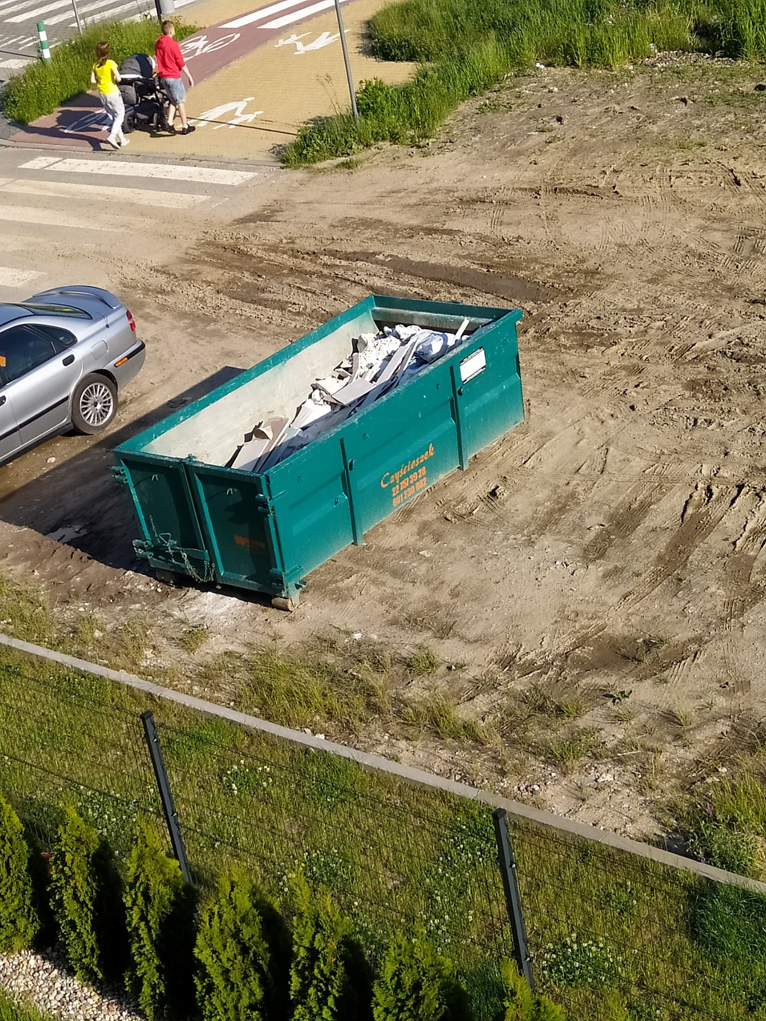Zielony kontener na gruz wypełniony odpadami budowlanymi, stoi na placu budowy z ubitym piaskiem, w tle para z wózkiem na przejściu dla pieszych i rowerzystów oraz zaparkowany samochód osobowy.