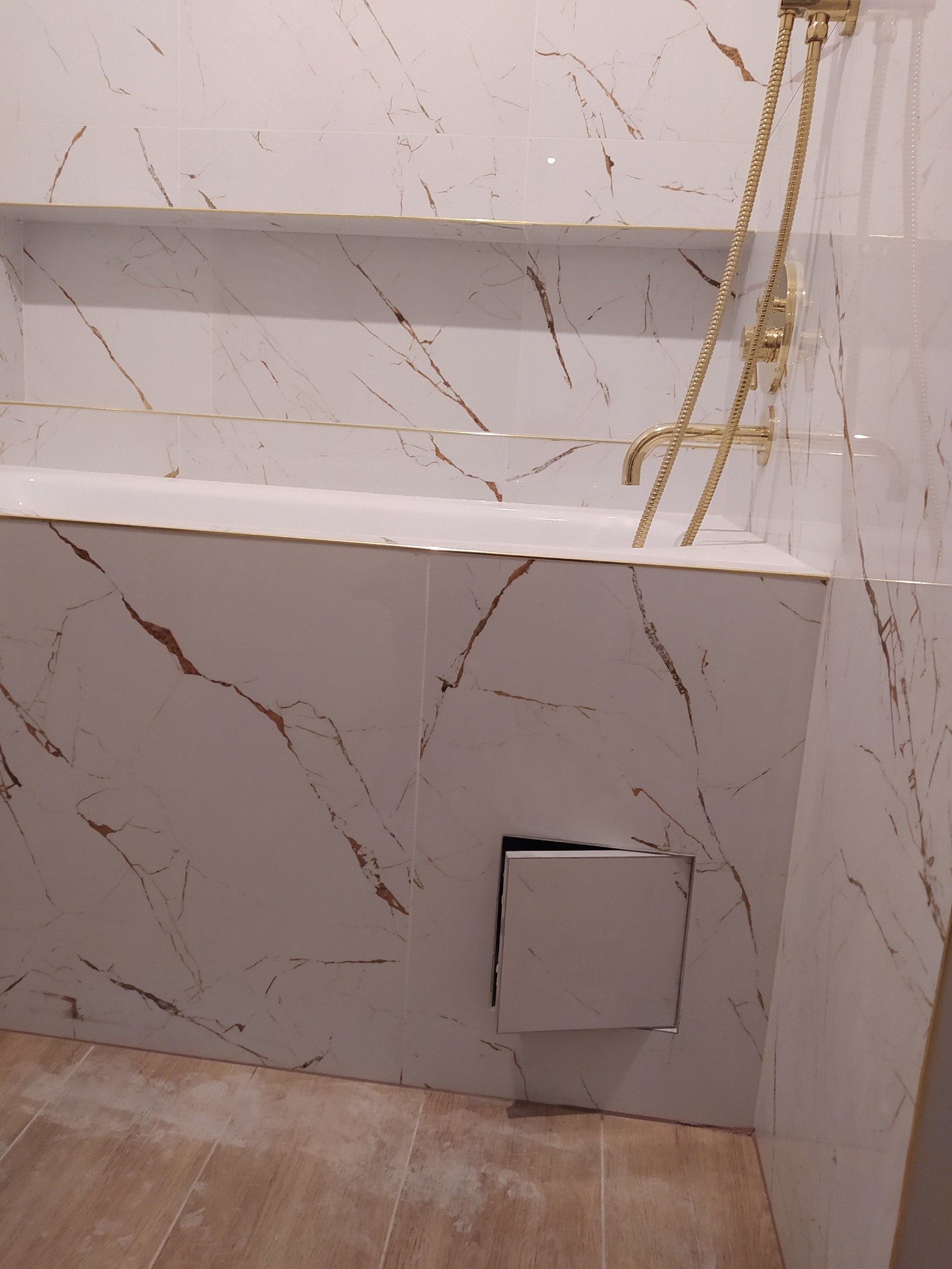 Wykończona łazienka z wanną obudowaną płytkami imitującymi marmur z złotymi żyłkami, złota bateria wannowa i prysznicowa, oraz ukryty rewizyjny właz instalacyjny.