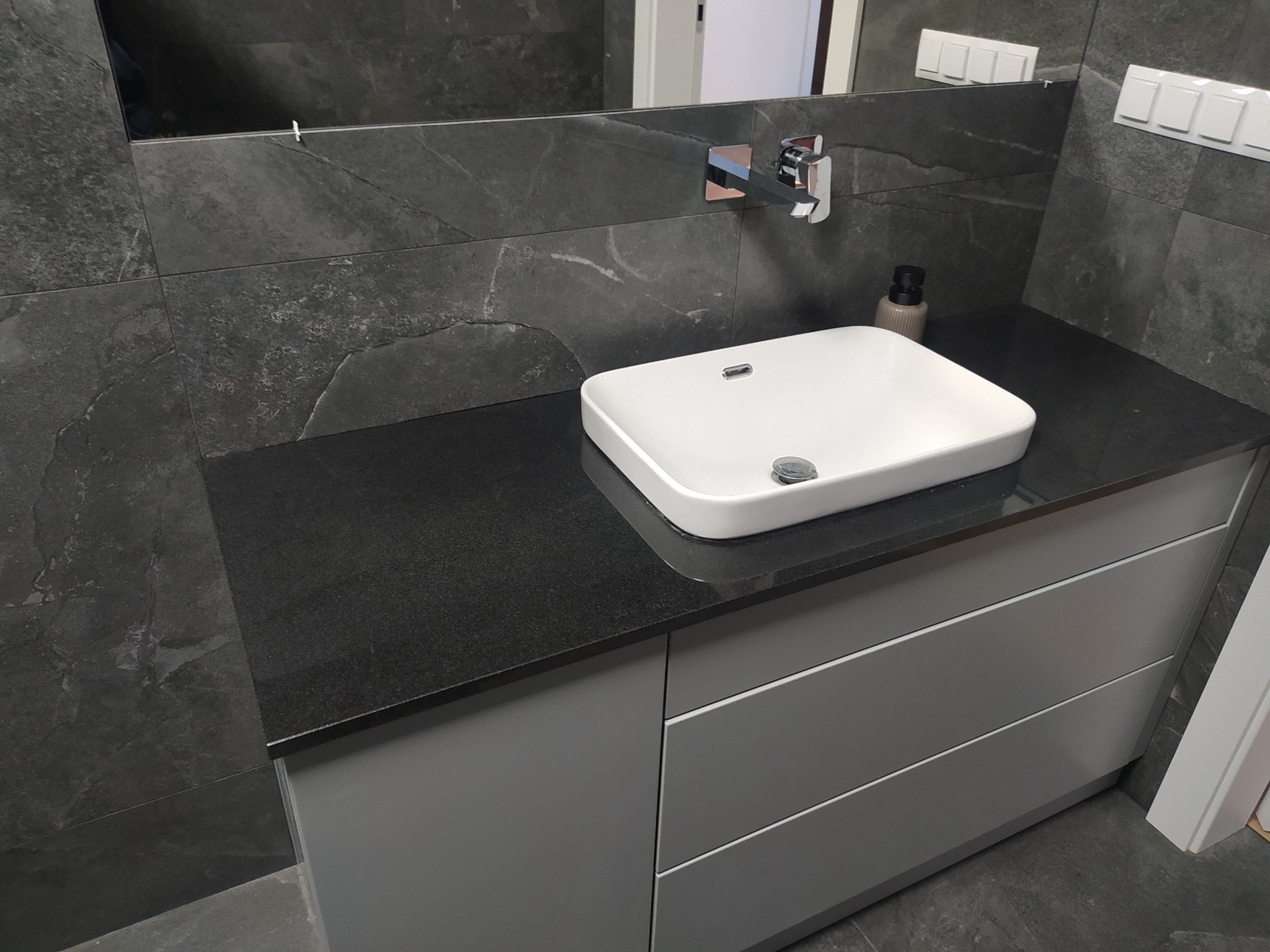 Nowoczesna łazienka z umywalką nablatową, ciemnym blatem i szarą zabudową szafkową z trzema szufladami, ściany wykończone ciemnymi płytkami imitującymi kamień.