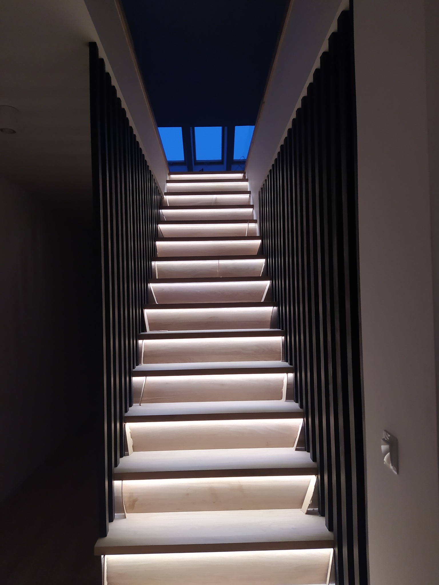 Nowoczesne schody drewniane z podświetleniem LED, czarne pionowe barierki, widok z dołu na świetlik dachowy.