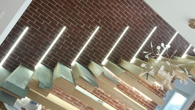 Skośne ujęcie sufitu z cegły klinkierowej z wpuszczonymi liniowymi oprawami LED, drewniane elementy konstrukcyjne i dekoracyjna lampa wisząca.