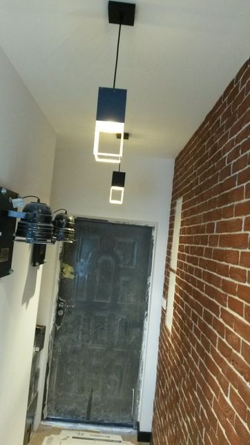 Nowoczesna lampa wisząca z kwadratowymi elementami, oświetlająca korytarz z ceglaną ścianą i czarnymi drzwiami, dodatkowe oświetlenie na ścianie po lewej stronie.