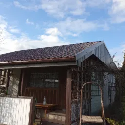 Niski dom z brązowym dachem z dachówki, drewnianą werandą i szarą elewacją z desek, na tle błękitnego nieba z chmurami.