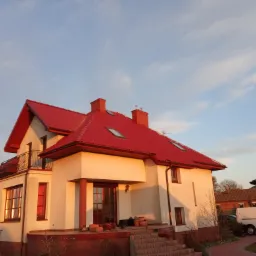Dwupiętrowy dom z jasną elewacją i intensywnie czerwoną dach&oacute;wką, widoczne okna dachowe, balkon z czarną balustradą, oświetlony promieniami zachodzącego słońca.
