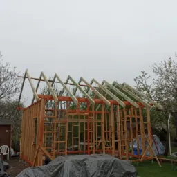 Konstrukcja szkieletowa budynku gospodarczego z widoczną więźbą dachową w trakcie montażu, na tle ogrodu z trampoliną i małym domkiem narzędziowym, pochmurne niebo.