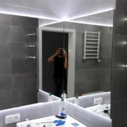 Wykończona łazienka z szarymi płytkami, podświetlanym lustrem i półkami. W lustrze odbija się osoba robiąca zdjęcie. Na blacie widoczne akcesoria i środki czystości.