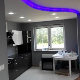 Nowoczesna kuchnia z szarymi ścianami, błyszczącą białą podłogą z żywicy i czarno-białymi szafkami, oświetlona listwą LED w kolorze fioletowym pod sufitem, z widocznym fragmentem drewnianej podłogi.