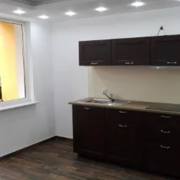 Nowoczesna kuchnia z ciemnymi, drewnianymi szafkami, jasnym blatem i płytkami na ścianie. Widok na balkon przez otwarte drzwi balkonowe.