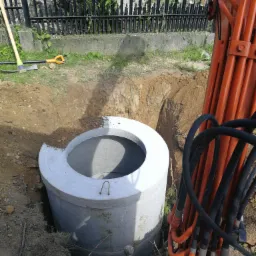 Betonowy krąg studzienny częściowo zagłębiony w wykopie, widoczne ramię koparki i narzędzia w tle.