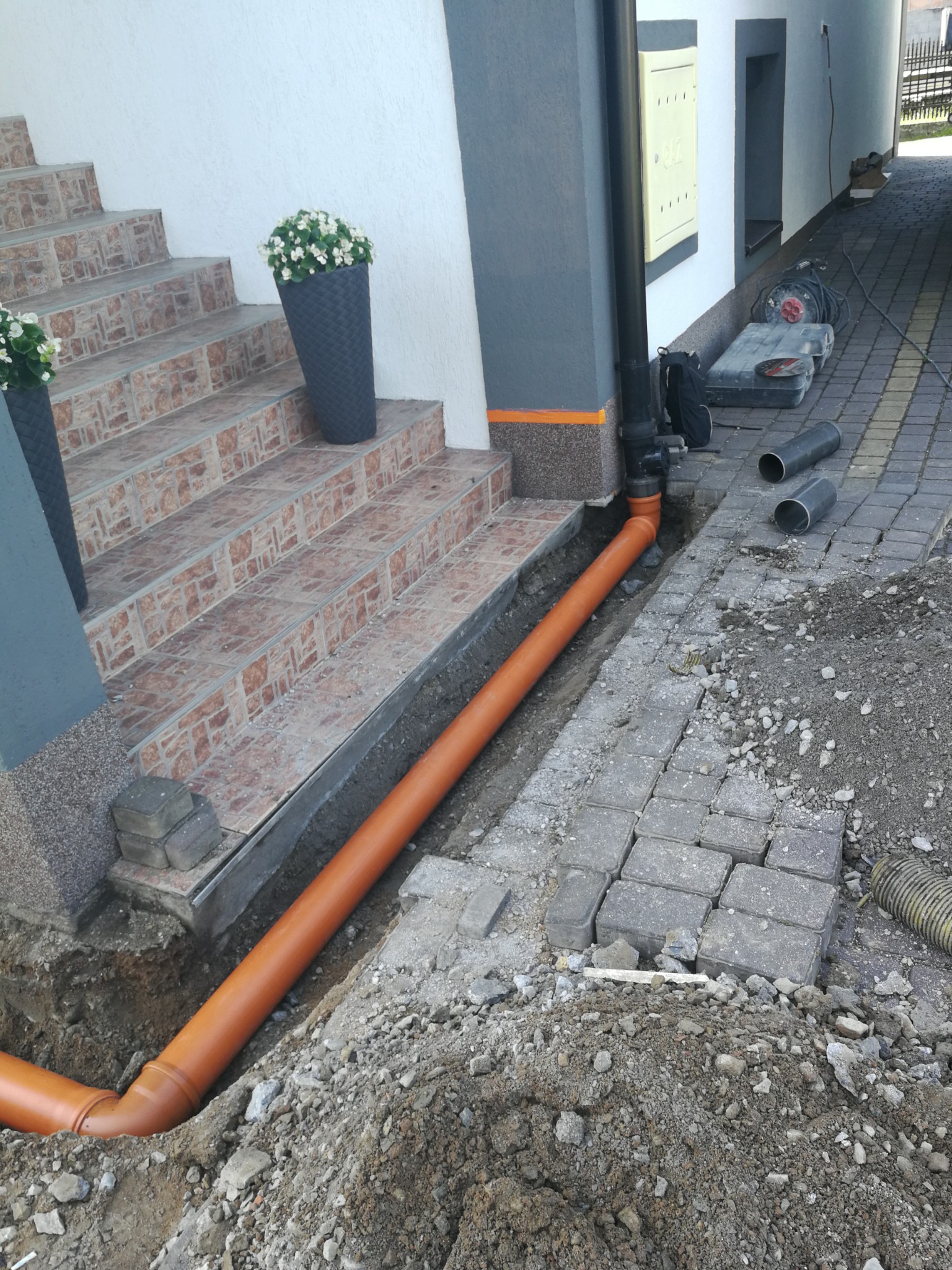 Pomarańczowa rura kanalizacyjna ułożona w wykopie obok schodów wejściowych do budynku, z widocznymi narzędziami i stertą ziemi.