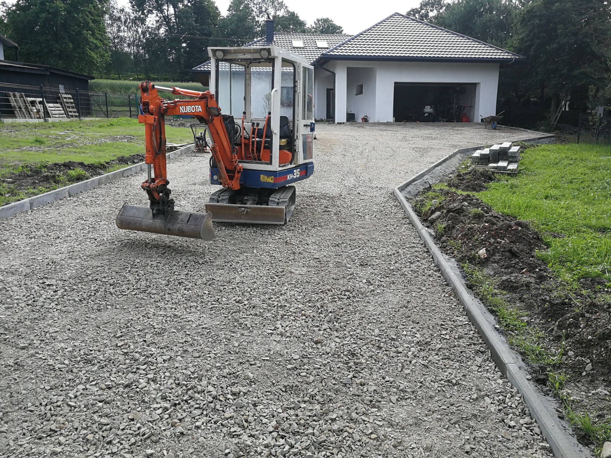 Pomarańczowy mini-koparka Kubota KH-35 na żwirowej nawierzchni przed białym domem z garażem, obrzeża chodnikowe po bokach, przygotowane do dalszych prac.