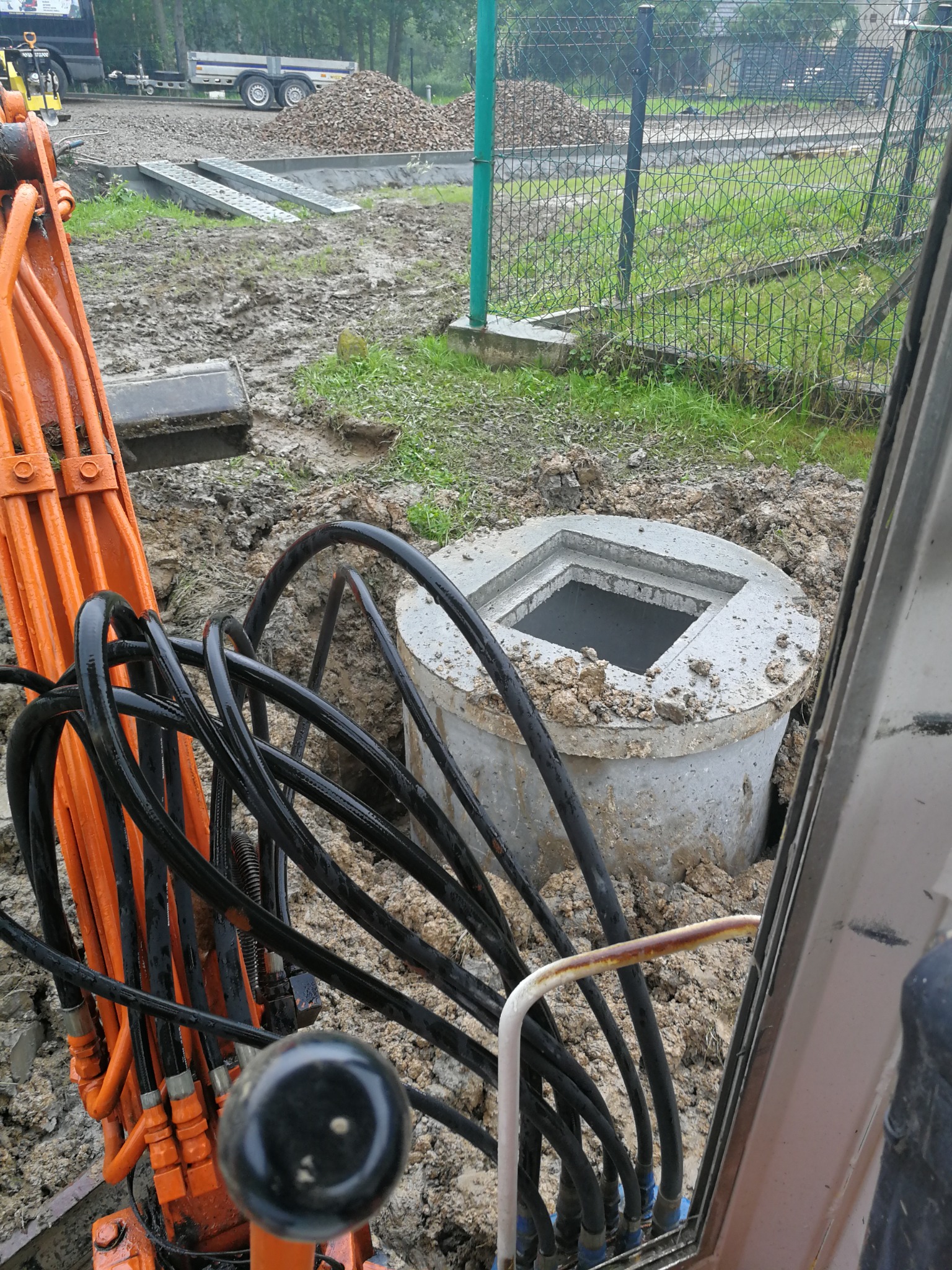 Betonowa studzienka z kwadratowym otworem w ziemi, widoczna z kabiny koparki, z pomarańczowymi przewodami hydraulicznymi i czarnymi wężami w pierwszym planie, w tle ogrodzenie z siatki i hałda ziemi.