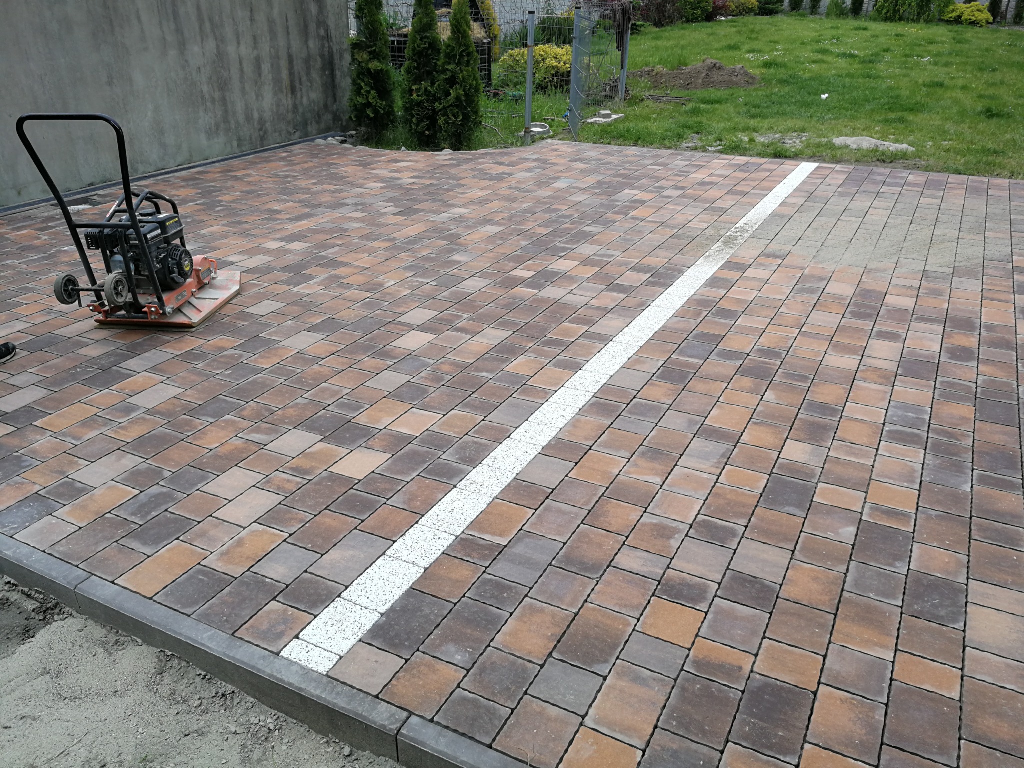 Układanie kostki brukowej z użyciem zagęszczarki w ogrodzie, widoczna linia z jasnej kostki oddzielająca obszary.