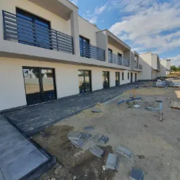 Nowoczesne domy w zabudowie szeregowej z ciemną kostką brukową na etapie układania, widoczne nieułożone kostki i piasek.