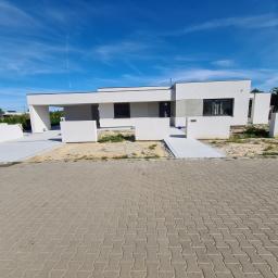 Damian Szulc Perfekt-Bud - Nowoczesny, parterowy dom z płaskim dachem, częściowo wykończony, z widocznymi elementami stanu surowego, na pierwszym planie ułożona kostka brukowa.